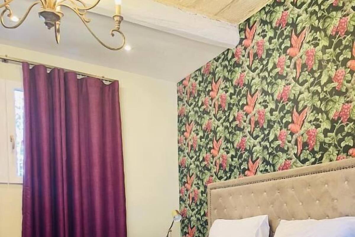 A neatly made bed with a purple throw blanket, a floral wallpaper, and a chandelier.