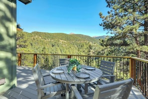Une terrasse en bois avec une table ronde dressée pour le repas, offrant une vue sur une chaîne de montagnes boisées.
