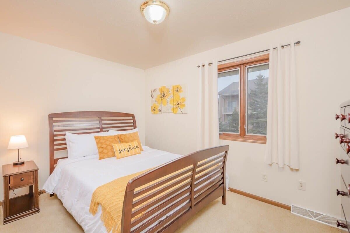 A bedroom with a wooden bed, a nightstand with a lamp, a window with curtains, and a wall with floral artwork.