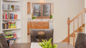 A dining area with a table, chairs, a bookshelf, and a kitchen in the background.