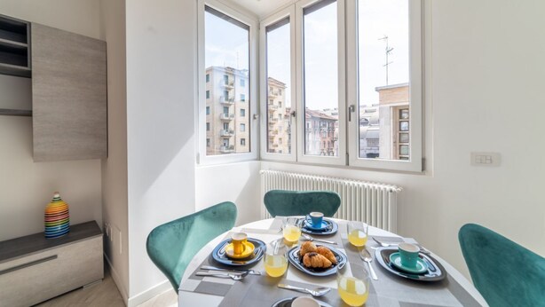 A dining area with a table set for breakfast, including croissants and orange juice, with a view of buildings through large windows. - Design Apartment - Central Station (Milano)