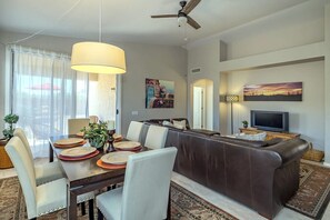 A modern living room with a sofa, dining table, and chairs. There is a television, a ceiling fan, and artwork on the walls. - Heatherglenn Velo - Comfortable Southwest Living (Oro Valley)
