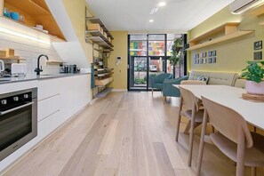 A modern kitchen with a built-in oven, a dining area with a table and chairs, and a view through a glass door to the outside.