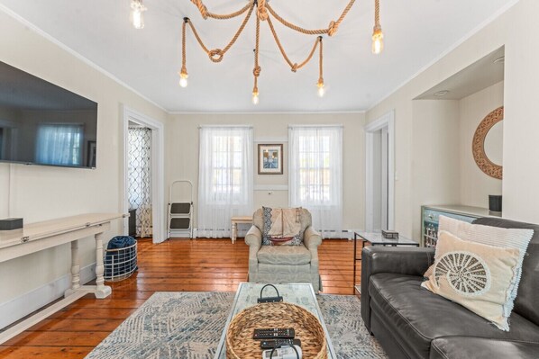 A spacious living room with a large flat-screen TV, a leather sofa, a coffee table, and a chandelier. - Historic 6-Bed Pet-Friendly Home, Walk to Seaport (Stonington)