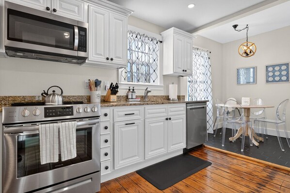 A modern kitchen with white cabinetry, stainless steel appliances, and a dining area with clear chairs and a wooden table. - Historic 6-Bed Pet-Friendly Home, Walk to Seaport (Stonington)