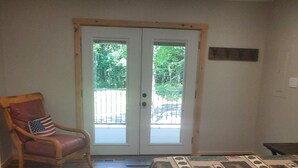 A room with a wooden floor, a bed with a patterned bedspread, a rocking chair with an American flag pillow, and a sliding glass door leading to an outdoor area.