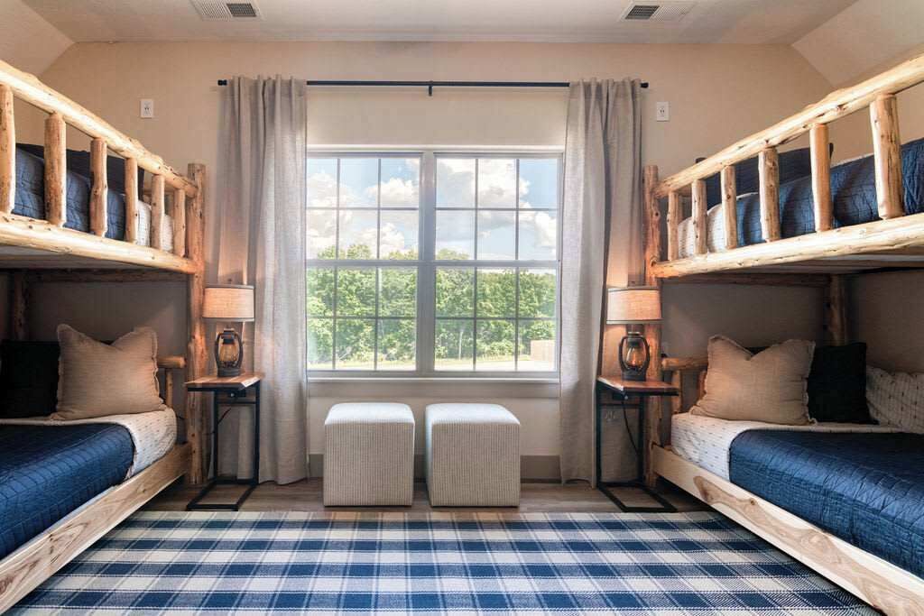 A room with two bunk beds, a window with curtains, a plaid floor, and bedside lamps.