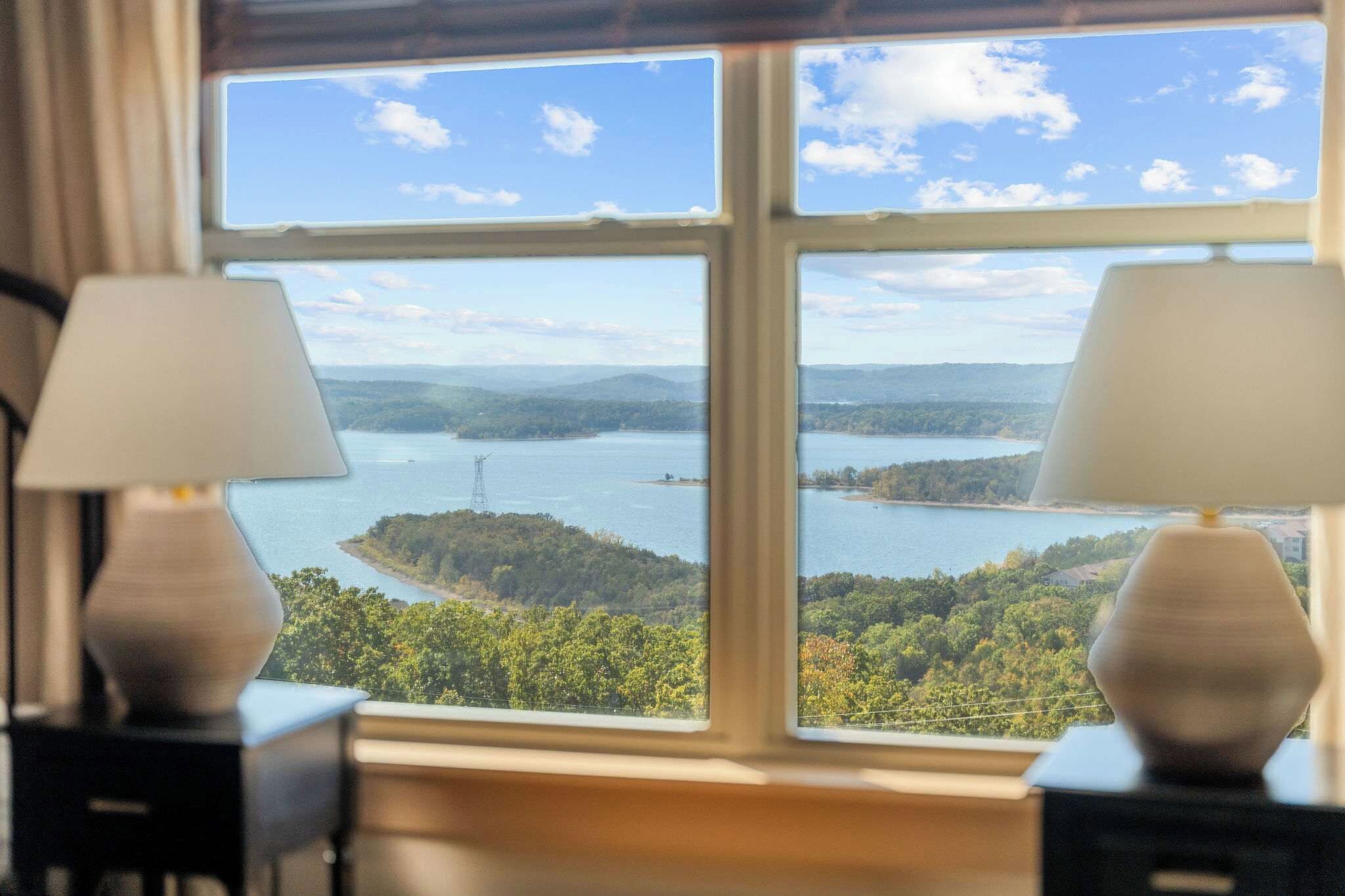 Et værelse med et stort vindue, der giver udsigt over en sø og det omkringliggende skovområde. Der er to lamper på hver side af vinduet.