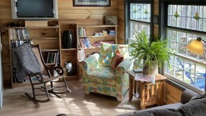 A cozy living room with a wooden interior, a rocking chair, a colorful armchair, a bookshelf, and a large window with a view. - The Pottery Cabin (Leicester)