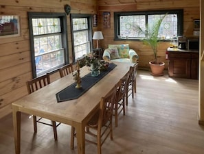 Een houten eettafel met stoelen, een bank en een bloempot in een kamer in chaletstijl, met houten wanden en grote ramen.