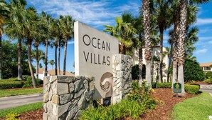 A sign for Ocean Villas with a stone base and a logo, surrounded by palm trees and a well-maintained lawn. - Ocean Villas #20 (St. Augustine)
