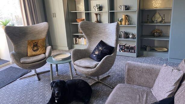 A modern living room with grey upholstered armchairs, a small round table, and a bookshelf filled with decorative items and books. A black dog is lying on the patterned carpet. - Stay in a grand old house, close to Hebden Bridge with parking & pet friendly.   (Hebden Bridge)