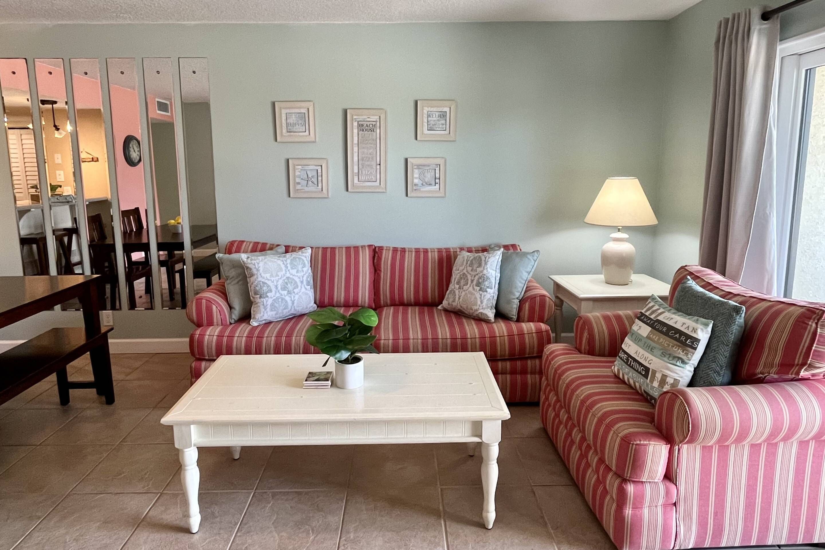 A living room with a striped sofa, a white coffee table, and a lamp. There are framed pictures on the wall and a plant on the coffee table.