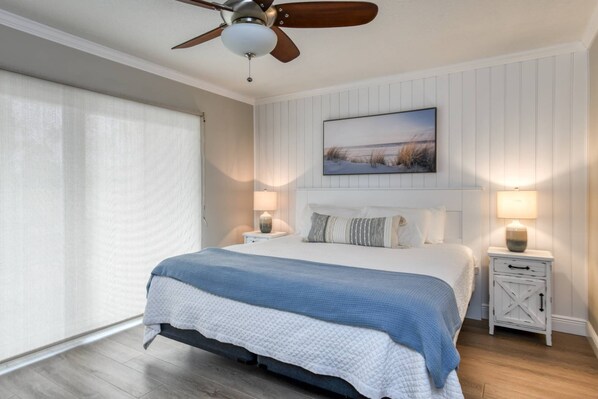 A bedroom with a bed, two bedside tables, a ceiling fan, a window with blinds, and a wall-mounted painting.