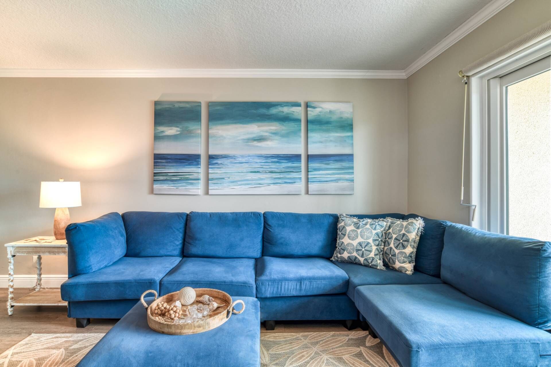 Un salon avec un canapé d’angle bleu, une petite table d’appoint avec une lampe et un mur orné de trois tableaux sur le thème de l’océan.