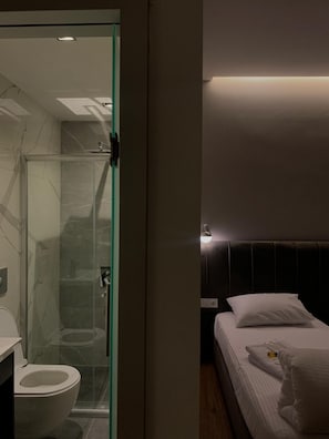 A modern bathroom with a glass shower enclosure, a toilet, and a bedside table with a lamp. - Ferah Hotel (Inegol)
