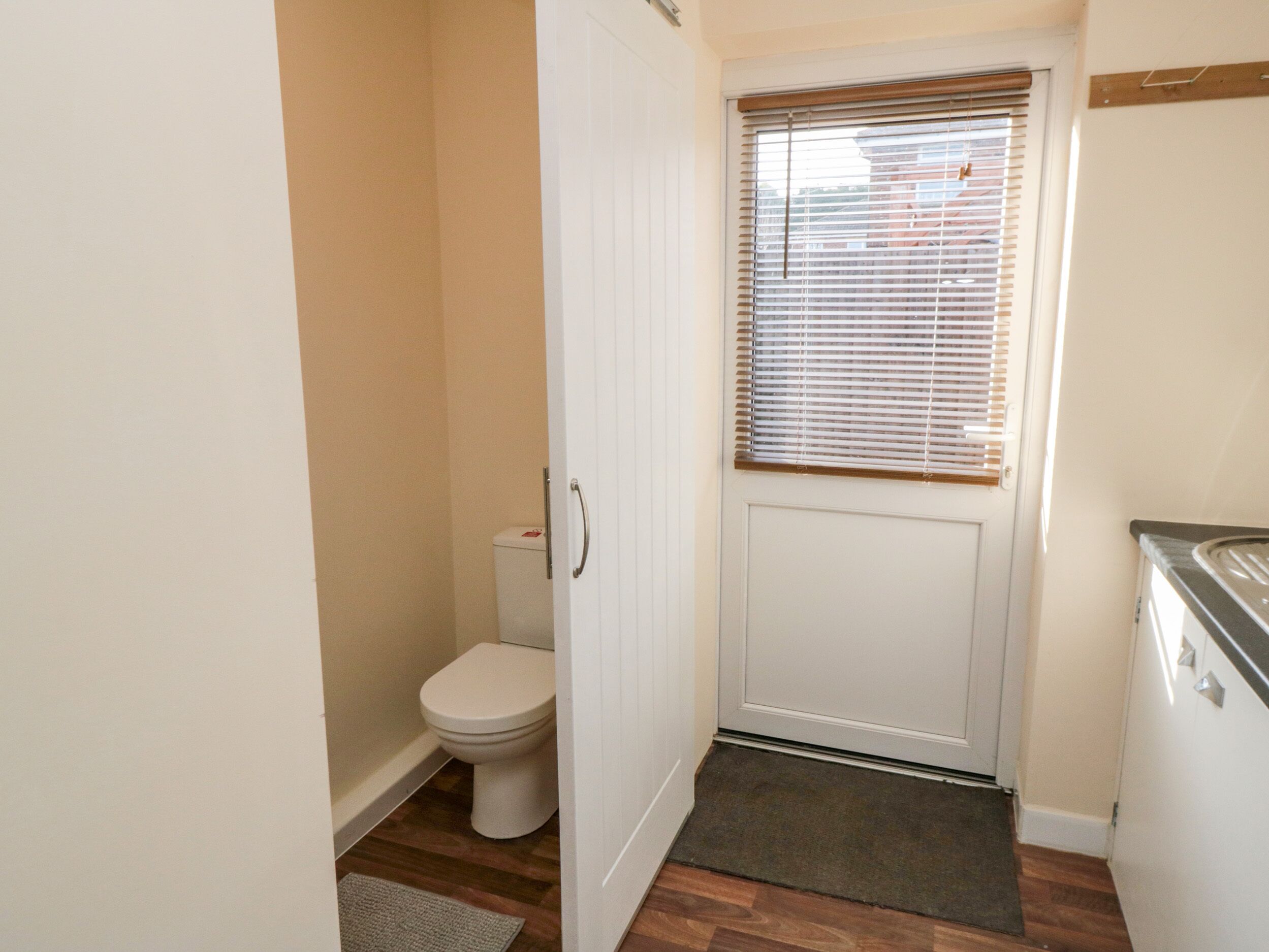 A small, narrow bathroom with a toilet, a door, and a window with blinds.