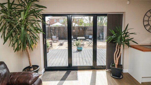 Una sala de estar con puerta corrediza de vidrio que da a un patio trasero. Hay dos macetas, un sofá de cuero marrón y piso de madera.