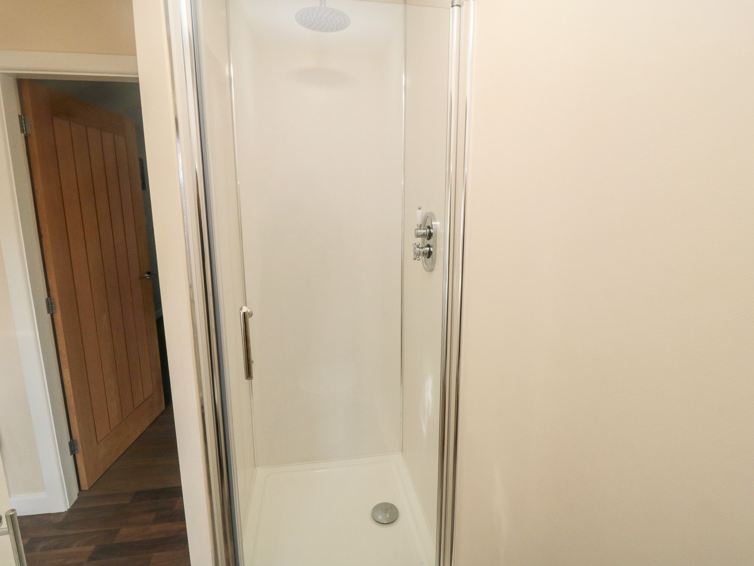 A glass-enclosed shower stall with a white floor, a door with a silver handle, and a wooden door visible in the background.