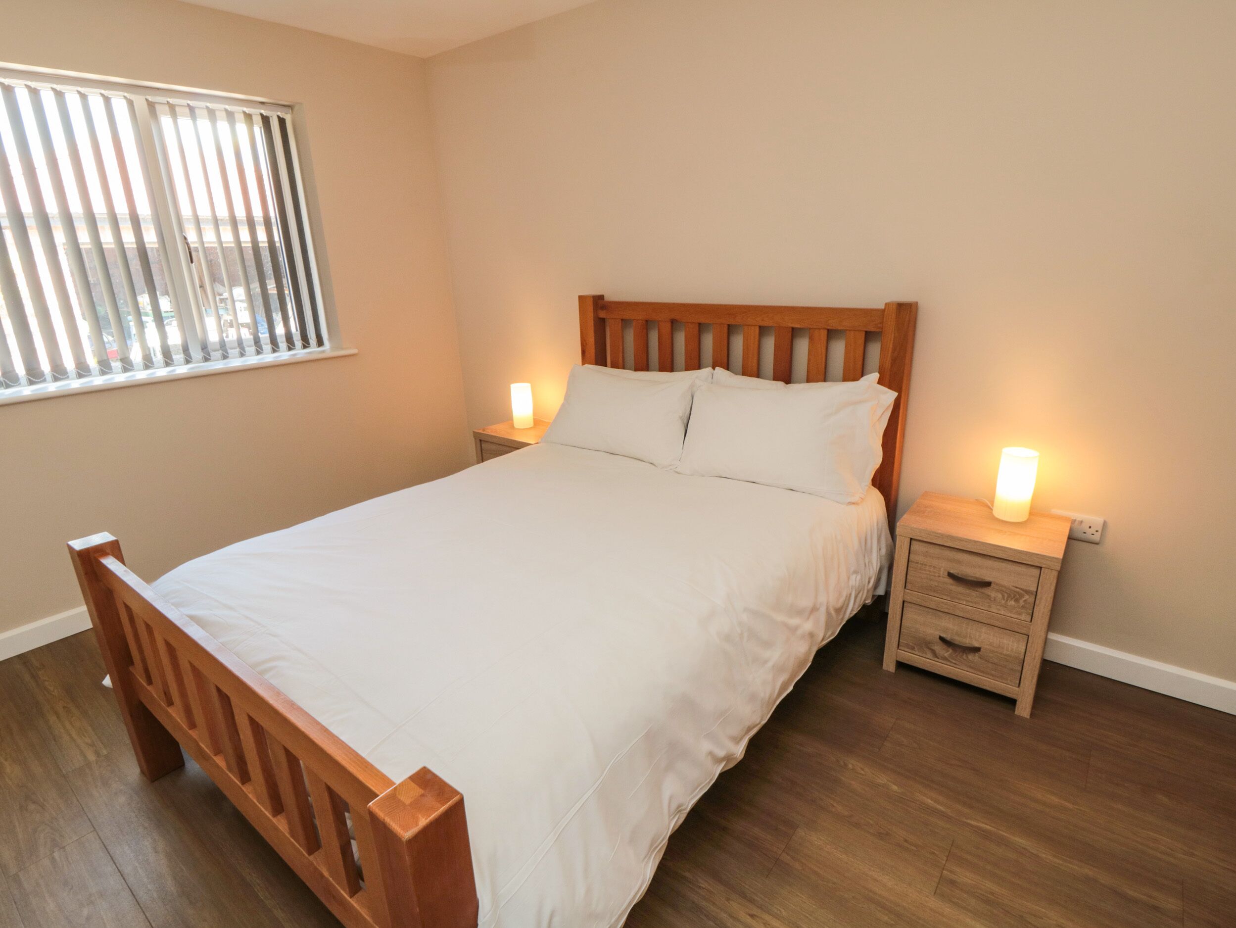 A neatly made bed with a wooden headboard, two bedside tables with lamps, and a window with blinds.