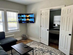 Ein Wohnzimmer mit Fernseher, einer Couch, einem Couchtisch und einem gemusterten Teppich. Zwei Türen führen in einen anderen Raum.