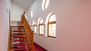 Un escalier avec des rampes en bois et des marches recouvertes d’une moquette rouge, menant à un palier doté de fenêtres en arc de cercle.