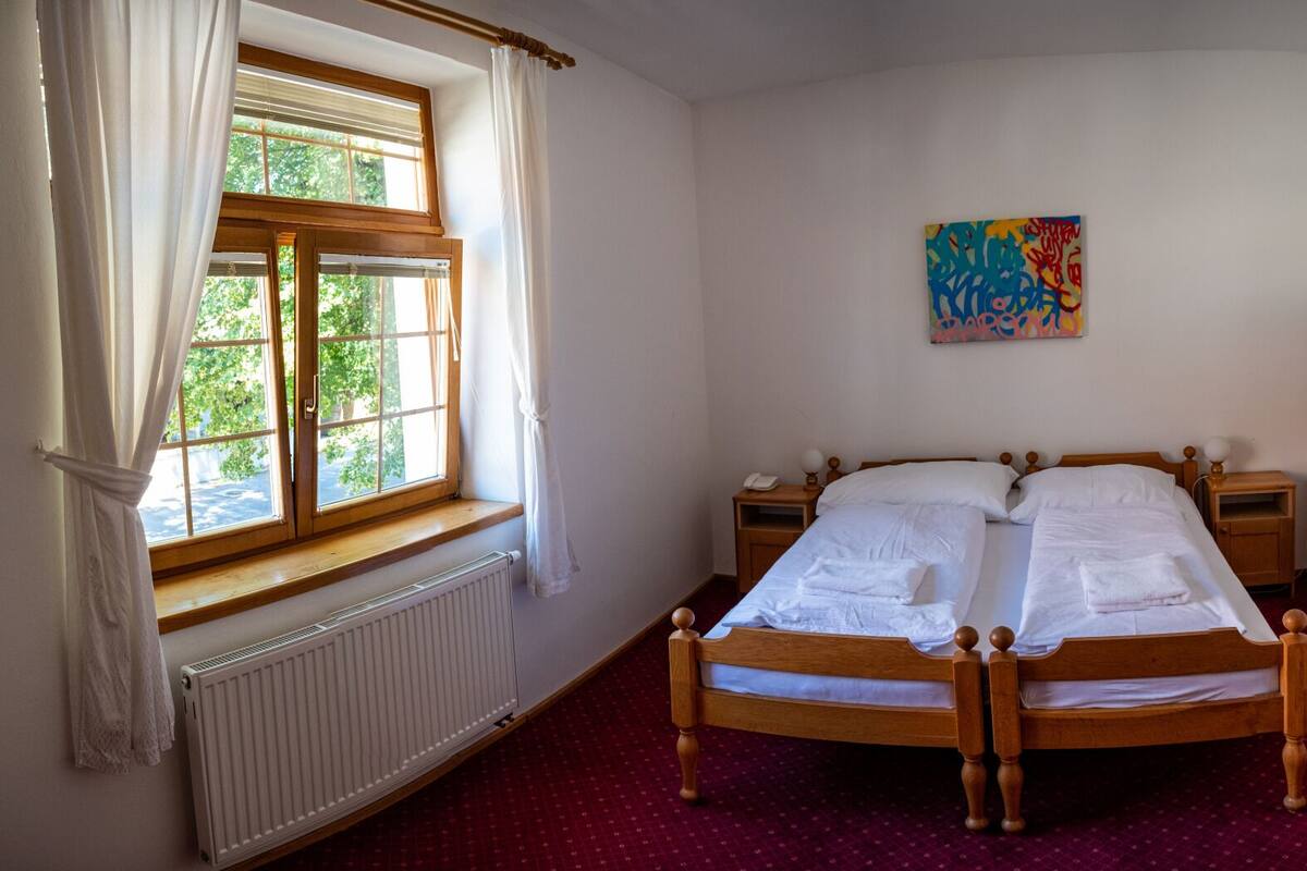 A bedroom with a double bed, a radiator, a window with curtains, a bedside table with a lamp, and a colorful painting on the wall.