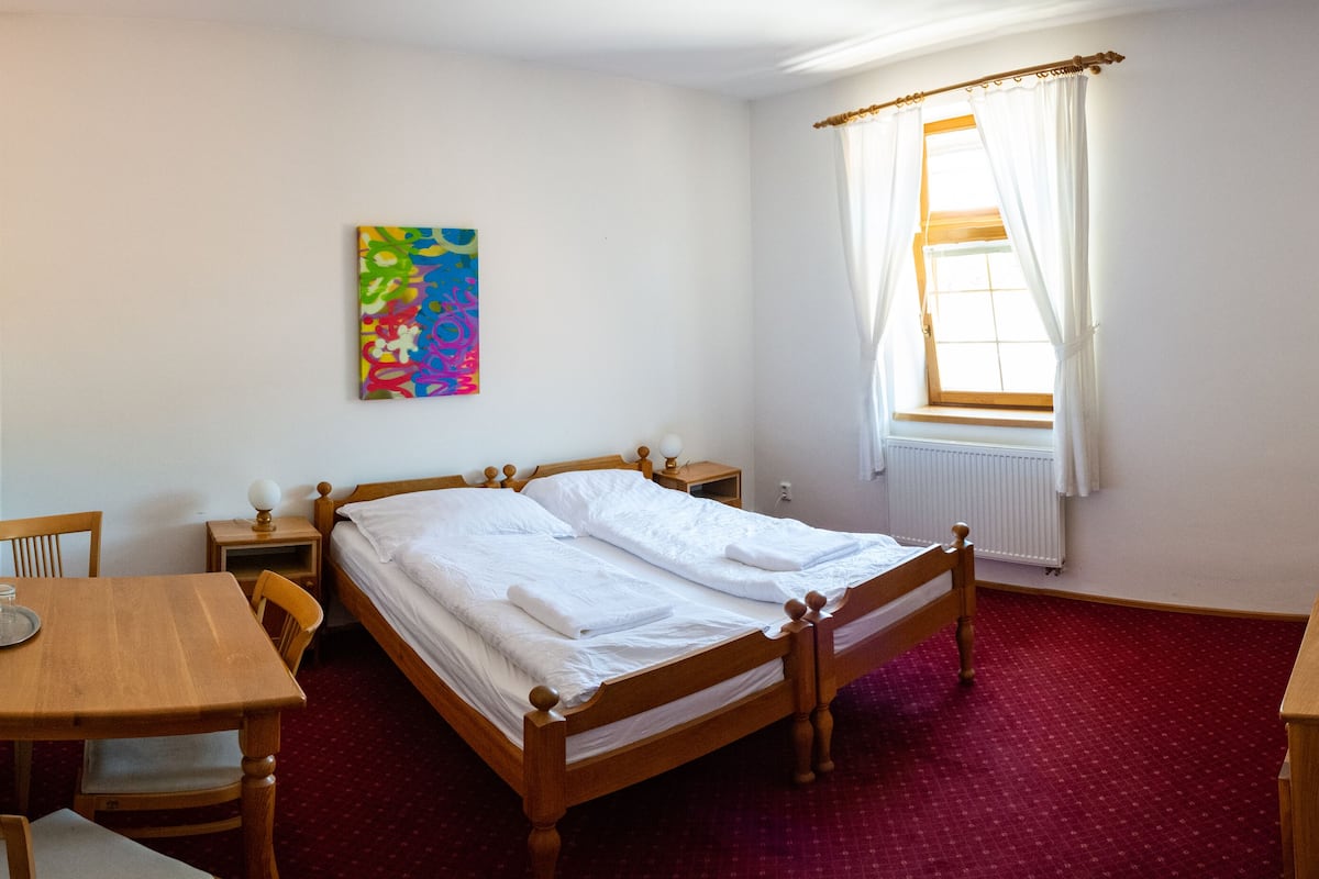 A bedroom with a wooden bed, a wooden desk, a chair, a bedside table, a radiator, a window with curtains, and a colorful abstract painting on the wall.