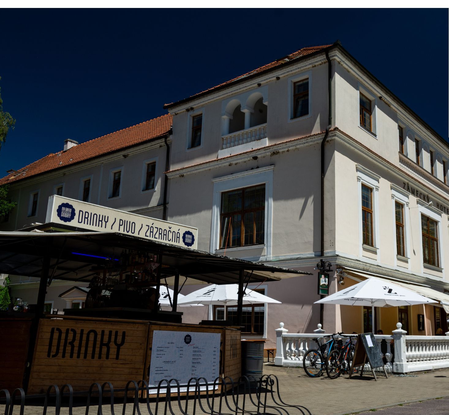 A two-story building with a sign for a restaurant named 'DRINKY / PIUO / ZAZRACNA'. There are outdoor umbrellas and a bicycle parked in front.