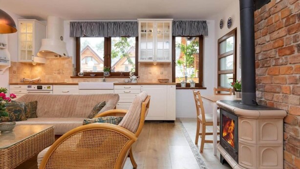 A cozy kitchen with a brick wall, white cabinets, a wooden dining table, wicker chairs, and a wood-burning stove.