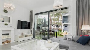 A modern living room with a glass coffee table, a grey sofa, a flat-screen TV, and a dining area visible through large glass doors. - Comfortable holiday home with garden, summer pool (Pobierowo)