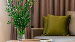 Una sala de estar con una mesa de centro redonda de madera, una planta verde en un florero de vidrio transparente, y un sofá gris con cojines verdes.