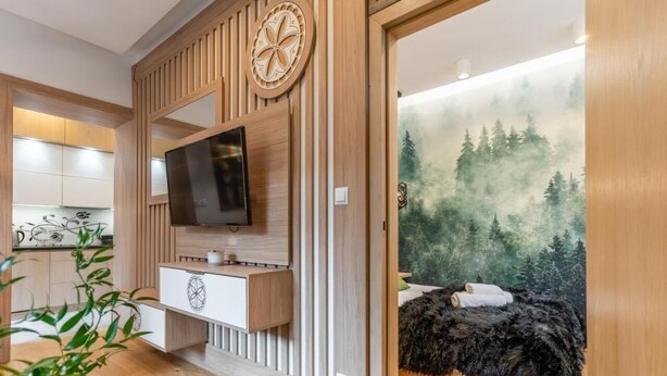 Un dormitorio moderno con una televisión instalada en un panel de pared de madera, una cama con un mural con motivos forestales y una mesita de noche de madera.