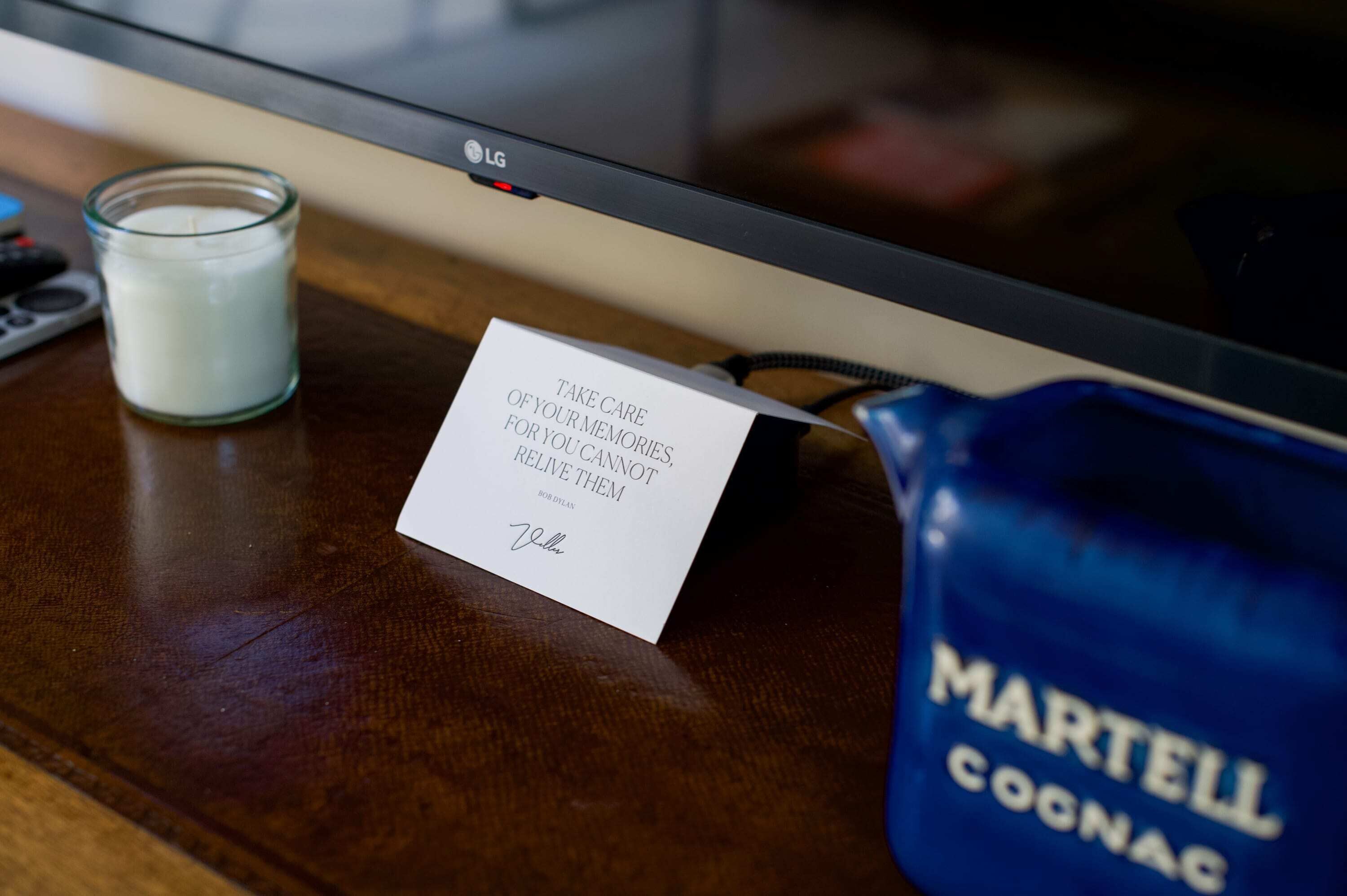 A flat-screen TV, a glass candle, a card with a quote, and a blue bag labeled 'Martell COCONAC' on a wooden surface.