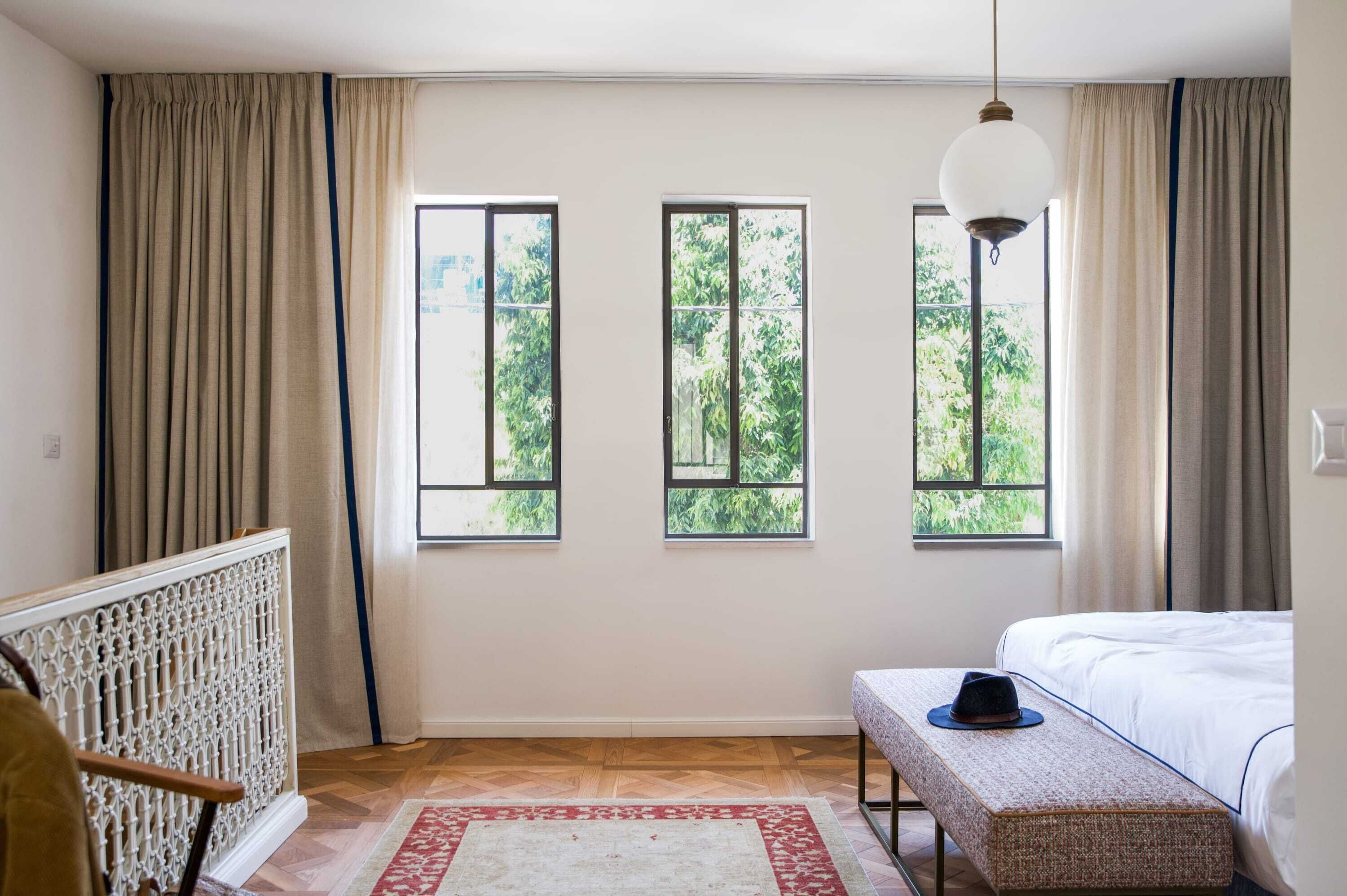 Ein Schlafzimmer mit einem Bett, einer Bank, einer Pendelleuchte und drei Fenstern mit Vorhängen.