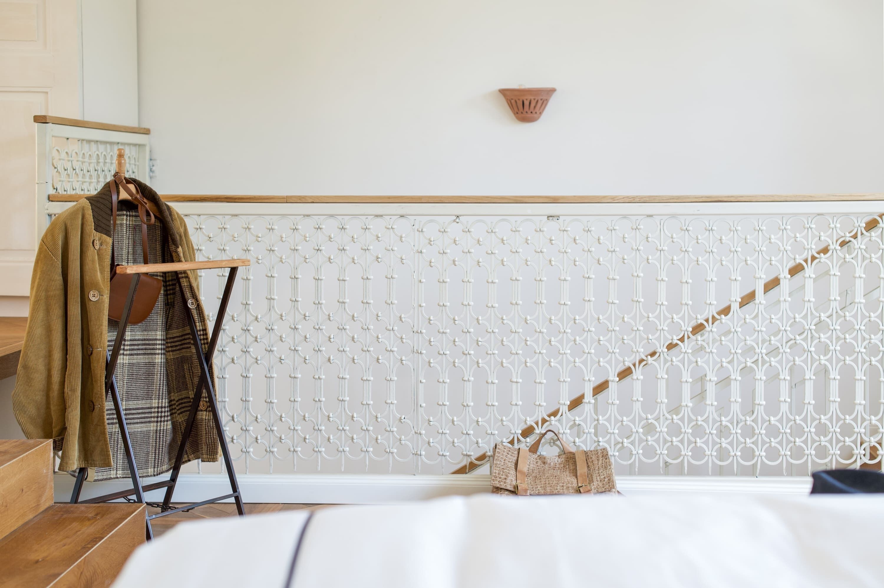 A room with a white patterned wall, a wooden floor, a bed with white bedding, a hanging rack with clothes, and a small woven bag.