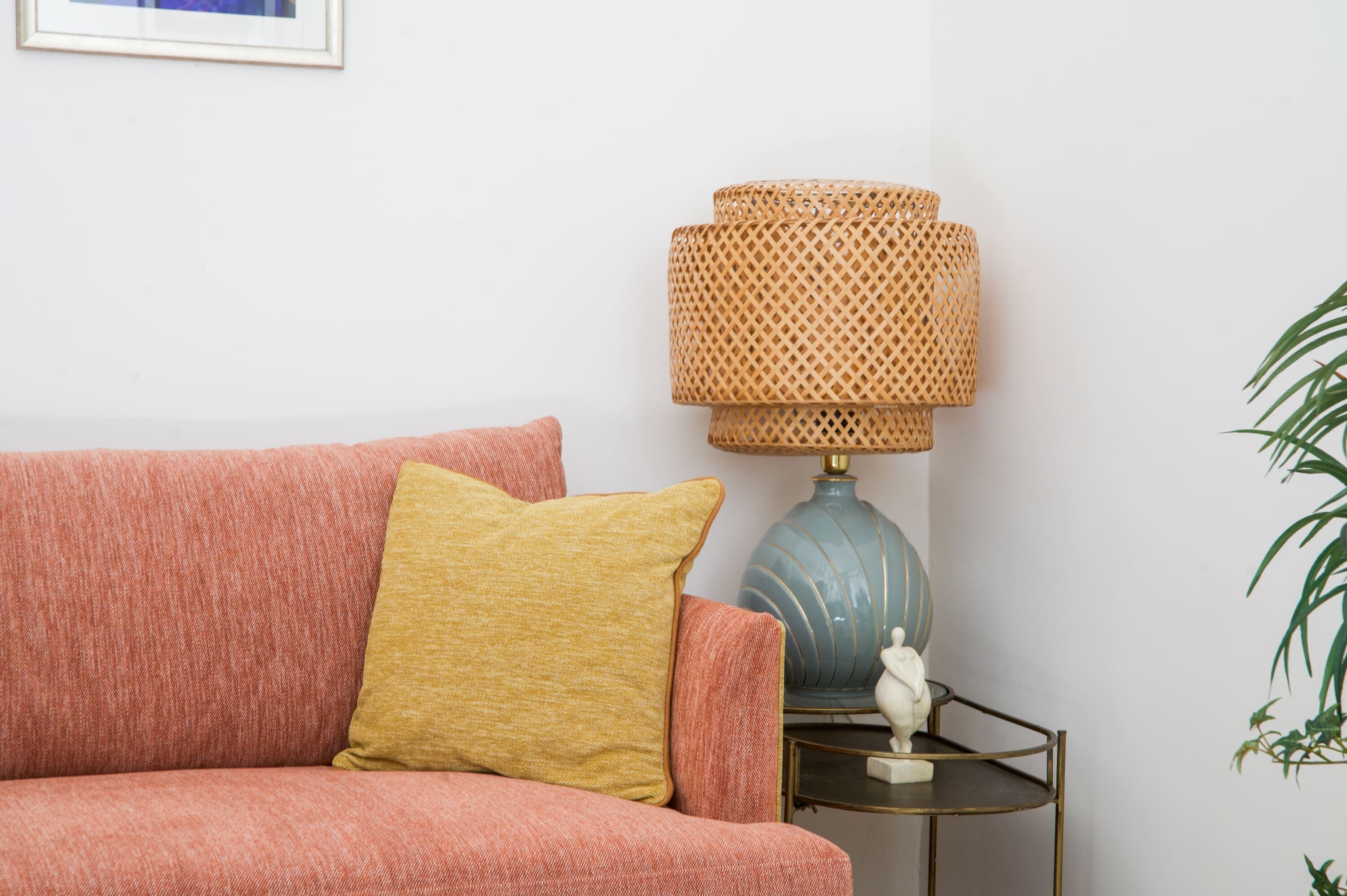 A pink sofa with a mustard-colored pillow, a blue lamp with a woven shade, and a small white statue on a metal side table.