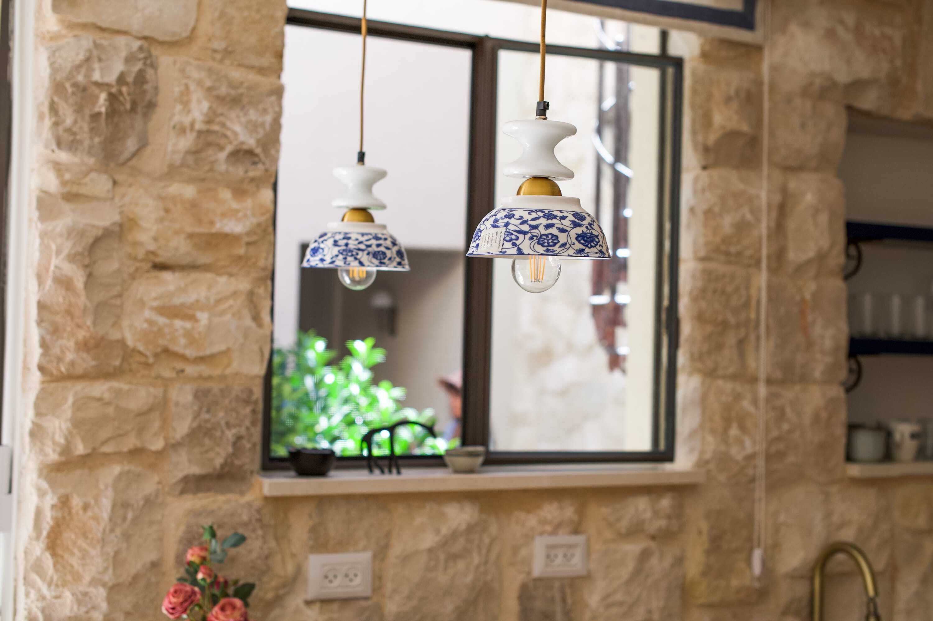 Une cuisine aux murs en pierre, une fenêtre donnant sur un paysage verdoyant et des suspensions lumineuses aux motifs bleus et blancs.