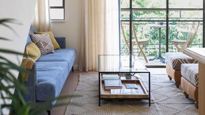 A living room with a blue sofa, a glass coffee table, and a view through large windows to an outdoor balcony. - Luxurious Urban Retreat with Private Outdoor Area (Tel Aviv-Yafo)