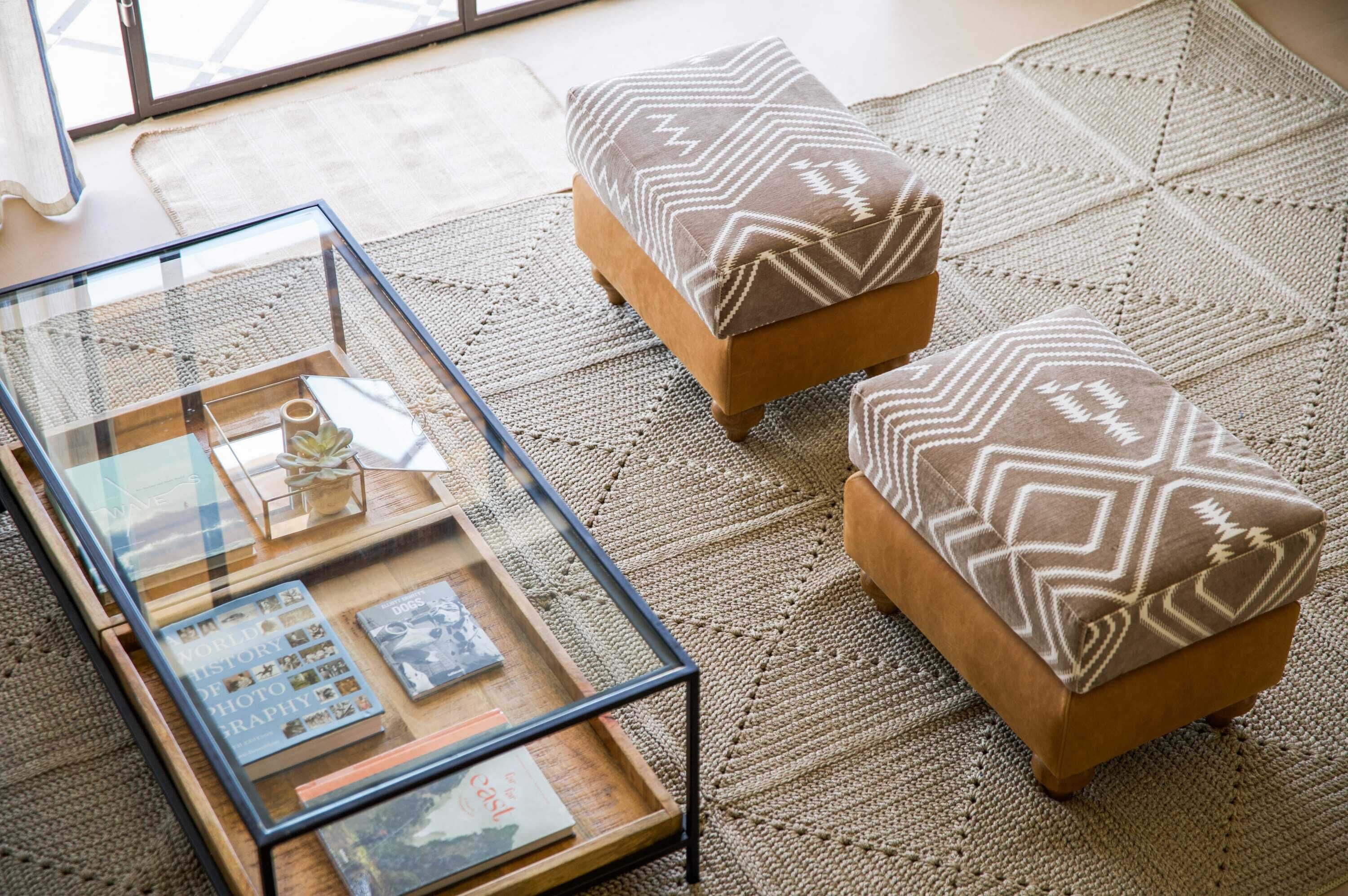 Un salon chaleureux avec une table basse en verre sur laquelle sont disposés des livres et des objets décoratifs, deux poufs à motifs et un tapis tissé.