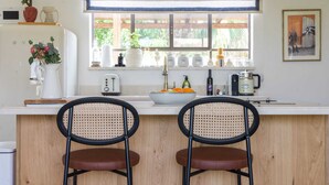 A kitchen with a white countertop, two wicker chairs, a window with a view of greenery, a refrigerator, and various kitchen items. - Luxurious Urban Retreat with Private Outdoor Area (Tel Aviv-Yafo)