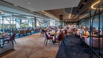 Een modern restaurant met een panoramisch uitzicht, met een mix van ronde en rechthoekige tafels en diverse zitmogelijkheden.