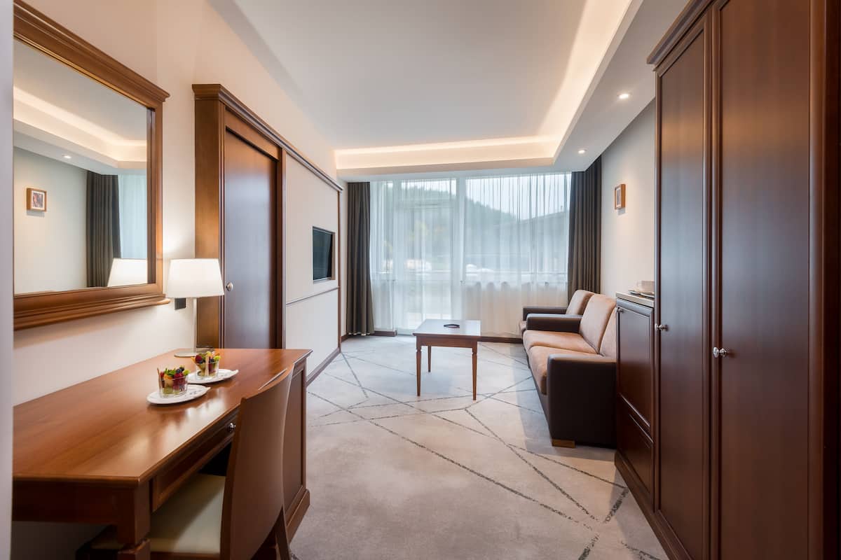 A hotel room with a wooden desk, a sofa, a coffee table, and a large window with curtains.