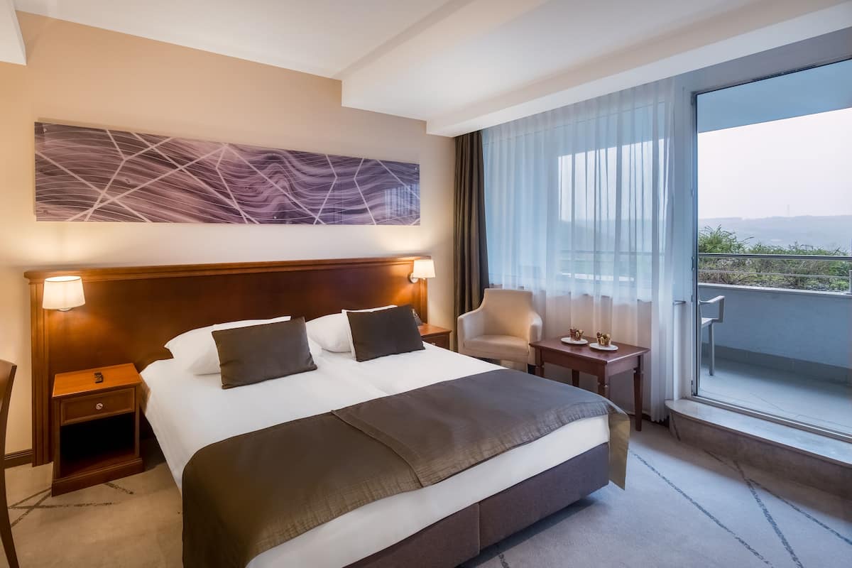 A hotel room with a large bed, two bedside tables with lamps, a chair, a small table with decorative items, and a view of the outdoors through large windows.