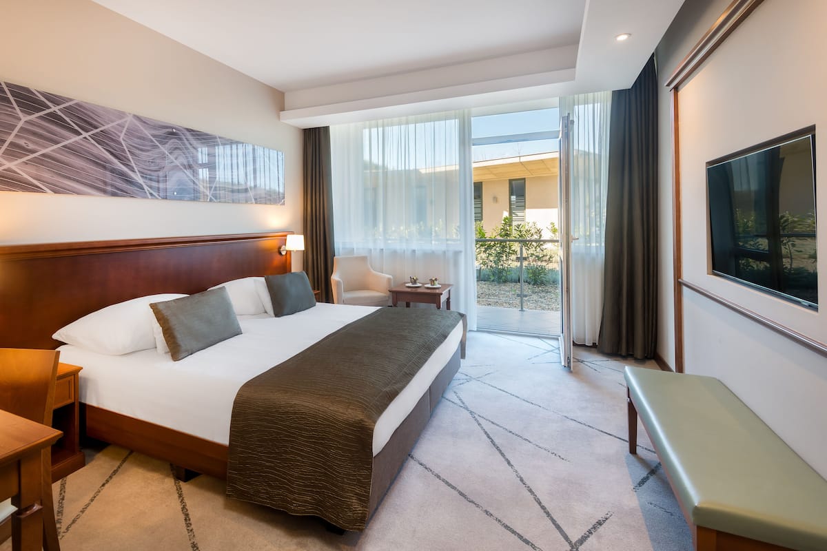 A hotel room with a large bed, a desk, a chair, a TV, and a view of the outside.