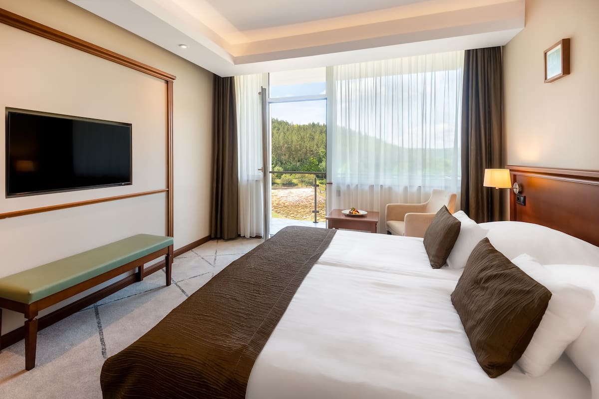 A hotel room with a large bed, a TV mounted on the wall, a bench, a desk with a lamp, and a view of the outdoors through the window.