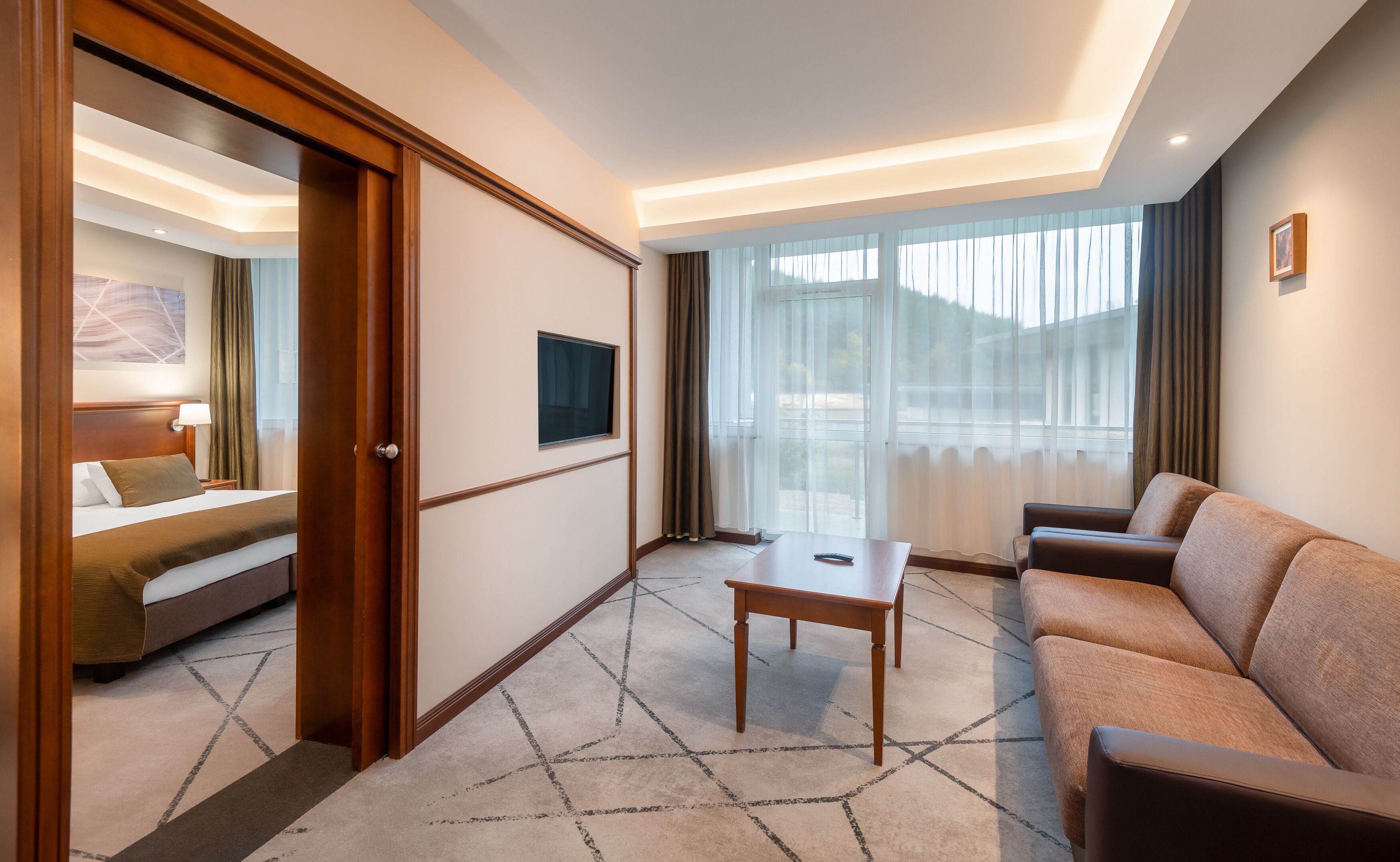 Habitación de hotel con cama, sofá, una mesita, televisor y un ventanal con cortinas.