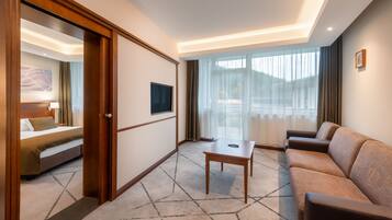 Habitación de hotel con cama, sofá, una mesita, televisor y un ventanal con cortinas.