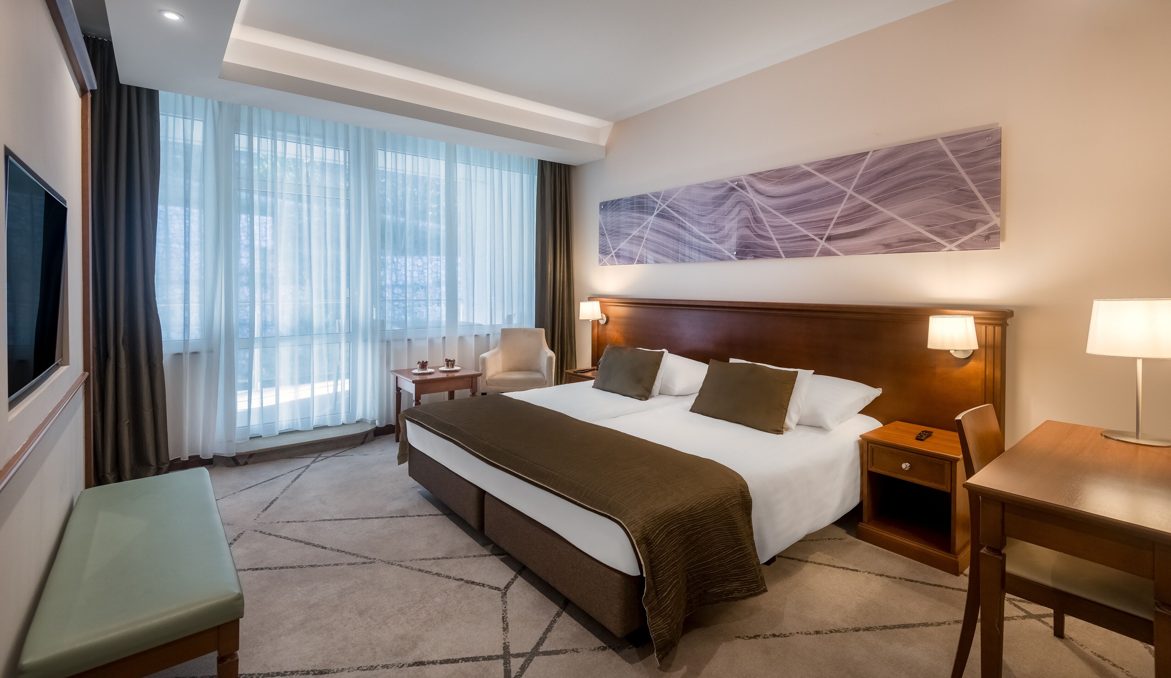 Ein Hotelzimmer mit einem großen Bett, einem Schreibtisch, einem Sessel, einem Fernseher und einem Fenster mit Vorhängen.