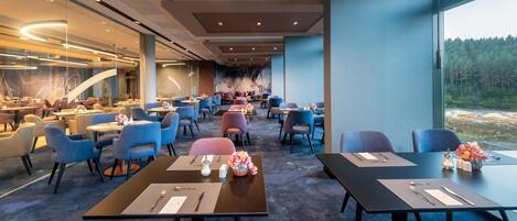 Un comedor de hotel moderno con varias mesas y sillas, un gran mural en la pared de fondo y vistas a un bosque a través de amplias ventanas.